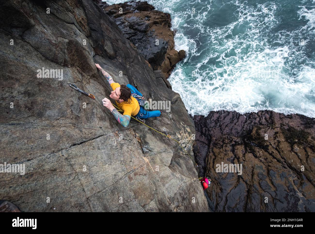 Ein Mann klettert auf eine Klippe am Meer und die Wellen schlagen darunter Stockfoto