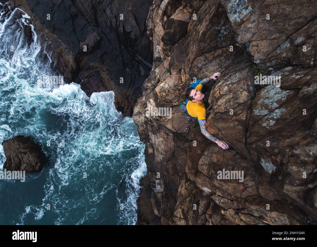 Ein Mann klettert auf eine Klippe am Meer und die Wellen schlagen darunter Stockfoto