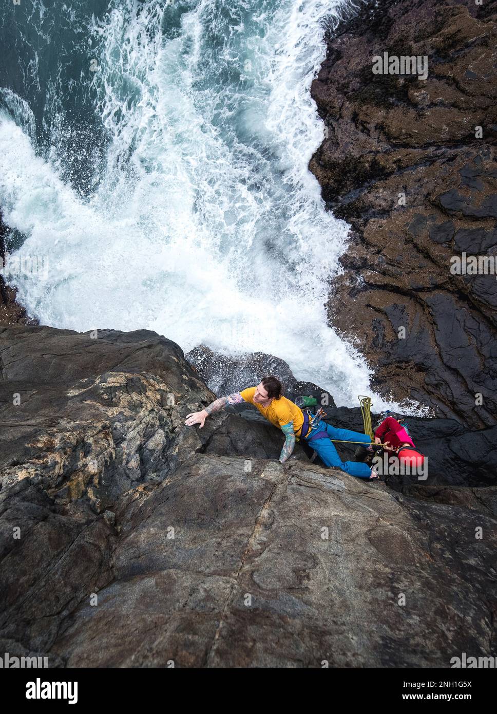 Ein Mann klettert auf eine Klippe am Meer und die Wellen schlagen darunter Stockfoto