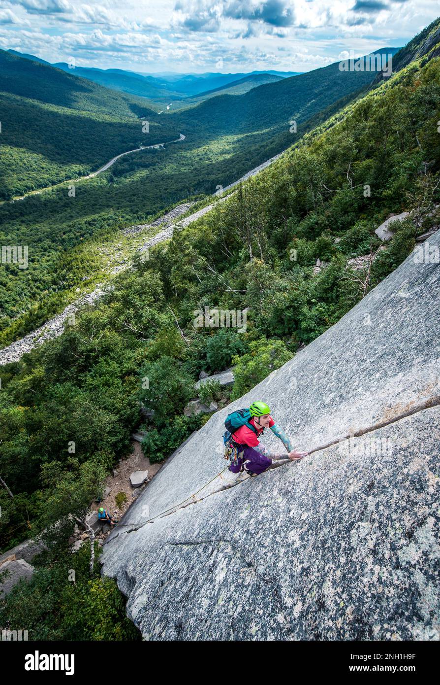 Ein Mann klettert steile Risse auf der Felswand mit dem Tal dahinter Stockfoto
