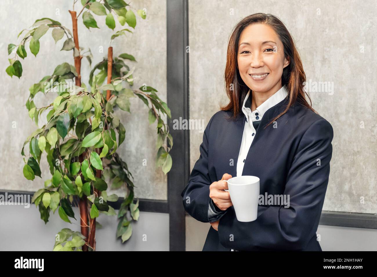Porträt einer positiven erwachsenen, ethnischen Unternehmerin im formellen Anzug, stehend mit einer Tasse heißes Getränk neben einem Topfbaum und schauend in die Kamera Stockfoto