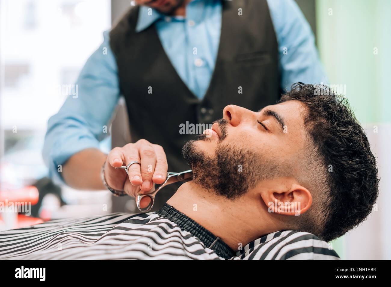 Schneiden Sie anonymen Barbier mit einer Schere, während Sie den Bart eines männlichen Klienten in einem professionellen Barbershop schneiden Stockfoto
