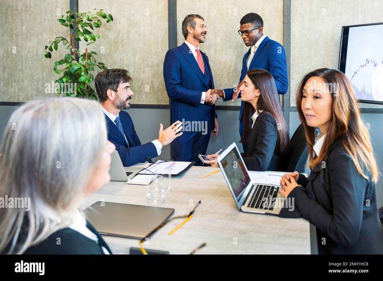 Eine Gruppe multiethnischer Kollegen versammelt sich im Sitzungssaal, während ein schwarzer Mann mit einer Kollegin die Hand schüttelt und eine asiatische Frau mit Laptop-Talki am Tisch sitzt Stockfoto