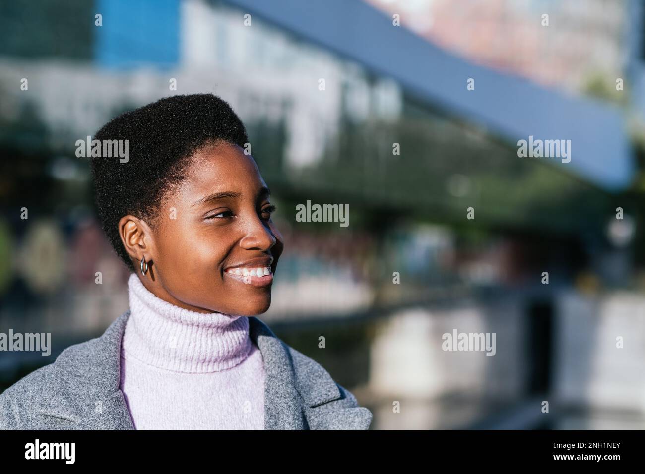 Optimistische schwarze Frau in stilvollem Pullover und Mantel mit kurzen Locken. Sie lächelt und blickt an sonnigen Tagen in der Stadt auf einen verschwommenen Straßenhintergrund Stockfoto