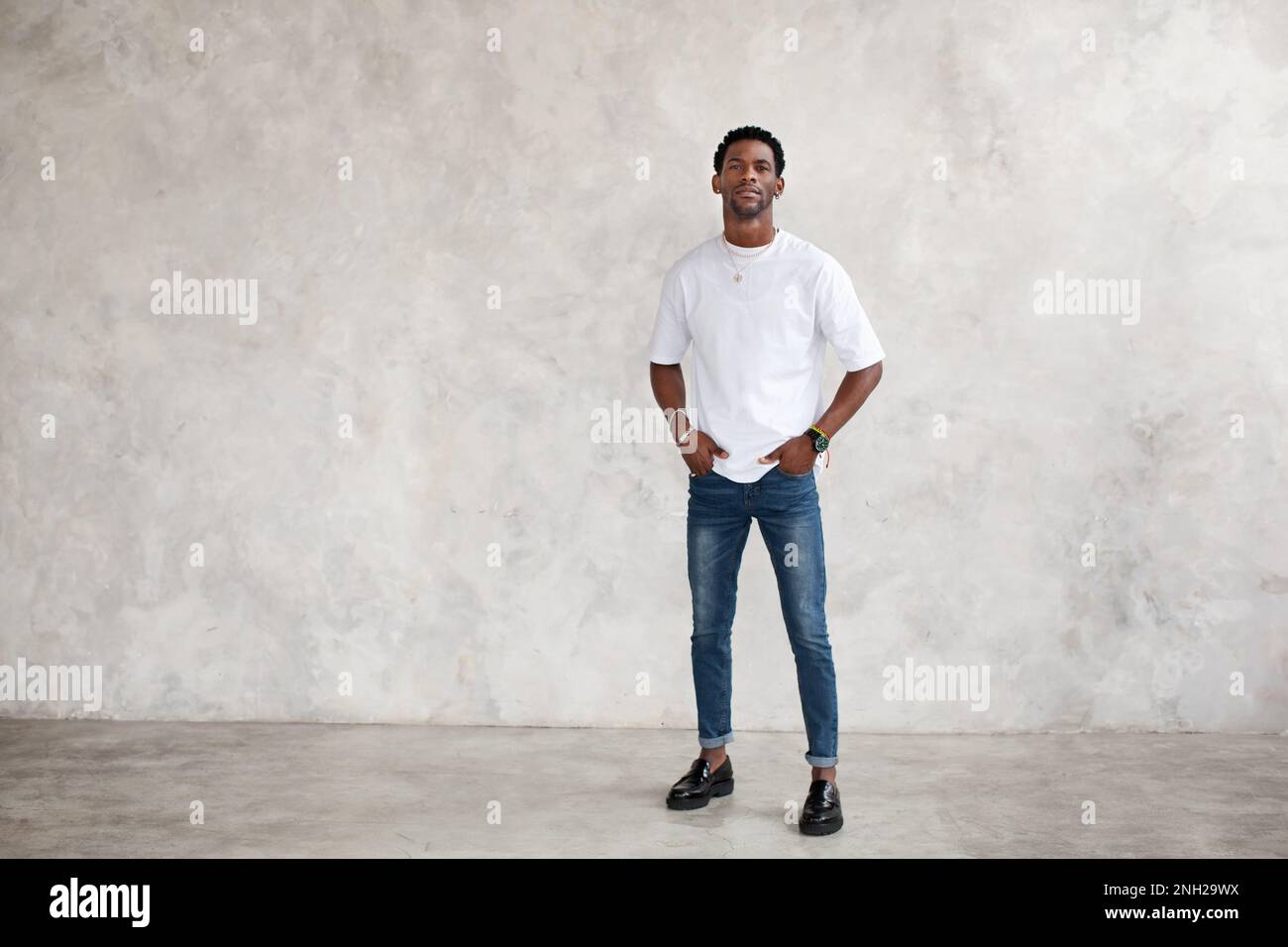 Ein afroamerikanischer junger Mann steht in voller Länge vor einer hellen texturierten Wand im Studio. Das männliche Model hat die Hände in den Taschen und trägt lässige Kleidung in Weiß Stockfoto