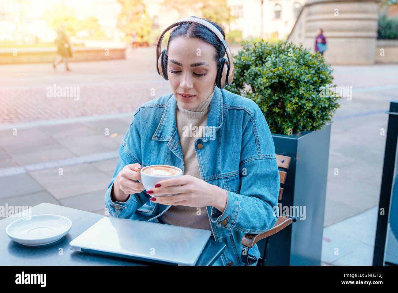 Frau in Jeansjacke und Kopfhörern trinkt Kaffee und hört Musik oder Hörbuch im Café. Lifestyle-Konzept Stockfoto