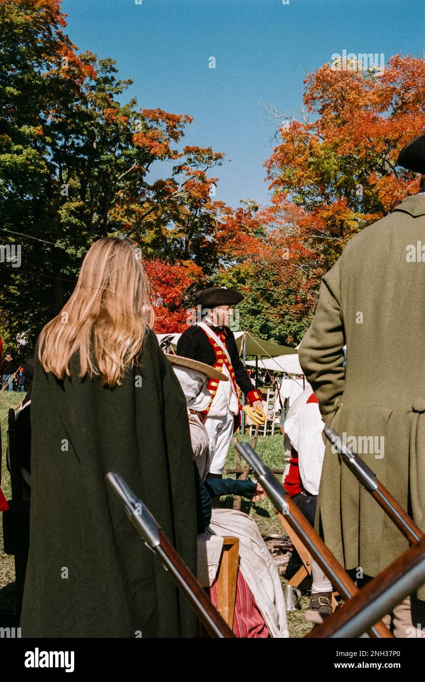 Ein amerikanischer Kolonialsoldat, der von einer Frau eingerahmt wird, folgt einem Soldaten und einem Ständer Musketen während der Nachstellung der Schlacht von Newbury im Unabhängigkeitskrieg. Die Stockfoto