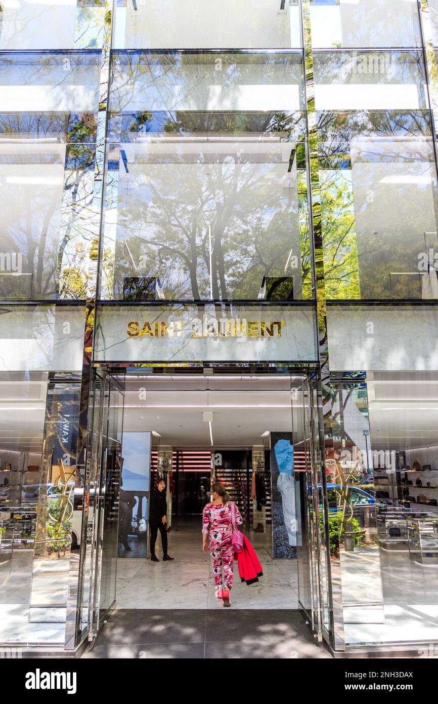 Französische modische Saint Laurent store in Tokyo in Omotesando. Tagsüber. Exterieur. Haupteingang Frau in Rosa, Rot und Weiß Hosenanzug wandern in. Stockfoto