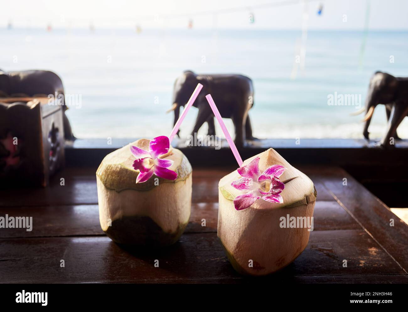 Zwei süße, frische Kokosnüsse mit Stroh und rosa Blüten im Restaurant mit hölzernen Elefanten am Strand mit Blick auf das Meer bei Sonnenaufgang. Stockfoto