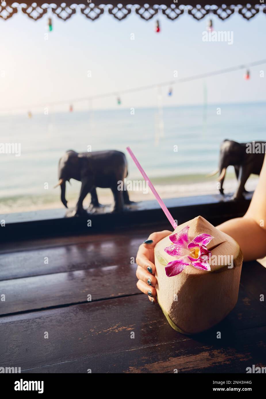 Frau trinkt süße frische Kokosnuss mit Stroh im Restaurant mit hölzernen Elefanten Dekoration am Strand mit Blick auf das Meer bei Sonnenaufgang. Stockfoto
