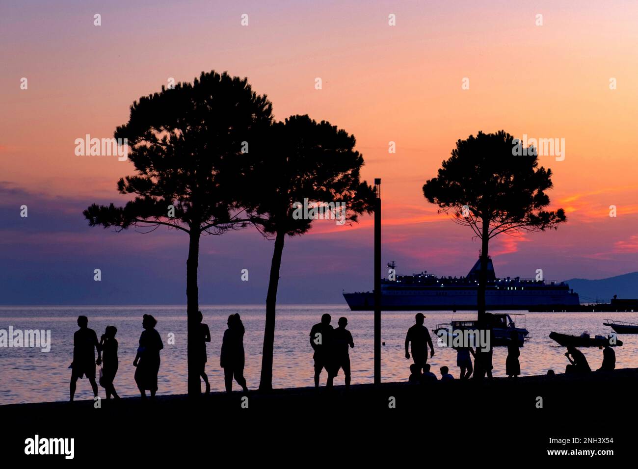 Touristen, die auf der Promenade spazieren gehen und Kreuzfahrtschiffe in der Bucht von Vlorë anlegen, die im Sommer bei Vlora im Südwesten Albaniens vor dem Sonnenuntergang abgeschirmt sind Stockfoto