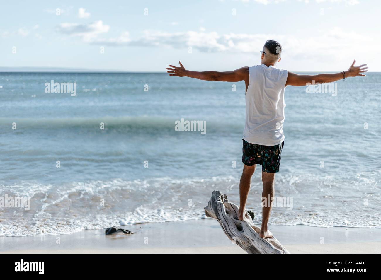 Ein Mann auf dem Rücken mit offenen Armen, der auf einem Baumstamm steht und den Sonnenuntergang am Strand beobachtet. Auf Reisen Stockfoto