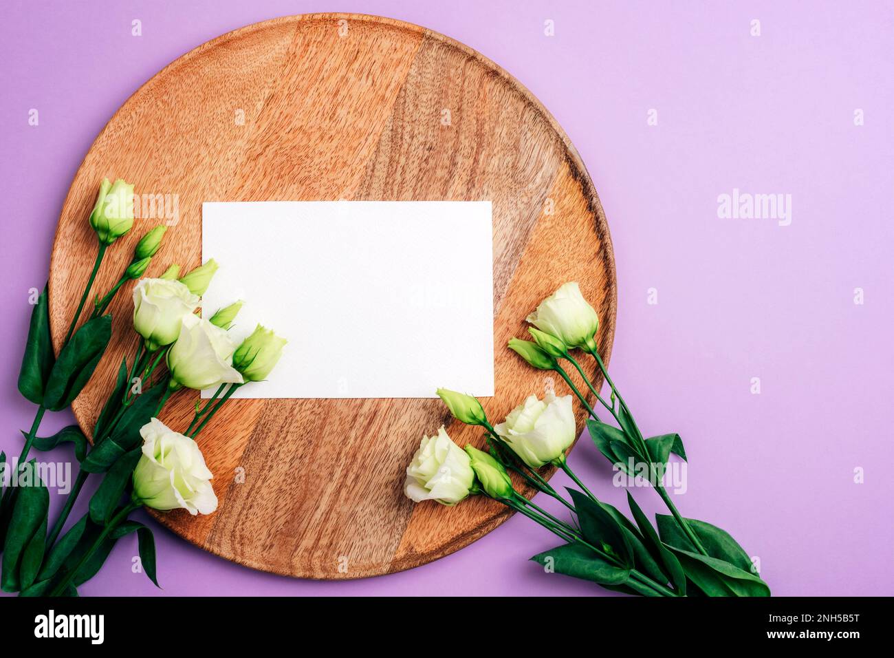 Eustoma grandiflorum weiße Blumen und leere Karte auf dem Holztablett auf lila Hintergrund, Modell. Festlicher Hintergrund, flach liegend, Kopierbereich. Stockfoto