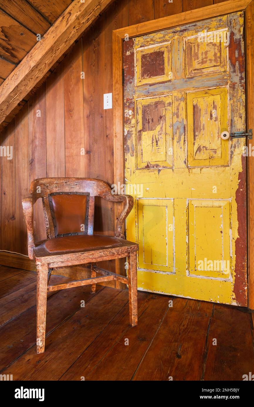 Gelb gestrichene Holzschranktür in Used-Optik, gepolsterter Holzsessel aus rötlichem Leder im oberen Schlafzimmer mit breitem Kiefernholzfußboden. Stockfoto