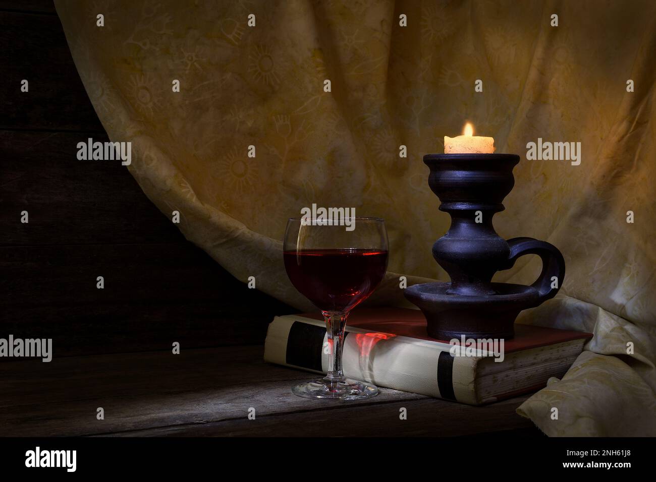 Ein klassisches Still Life-Ambiente mit einem Rotweinglas, einem alten Buch und einer beleuchteten Kerze in sanfter, warmer Stimmungsbeleuchtung mit einem Lichtschacht von links Stockfoto