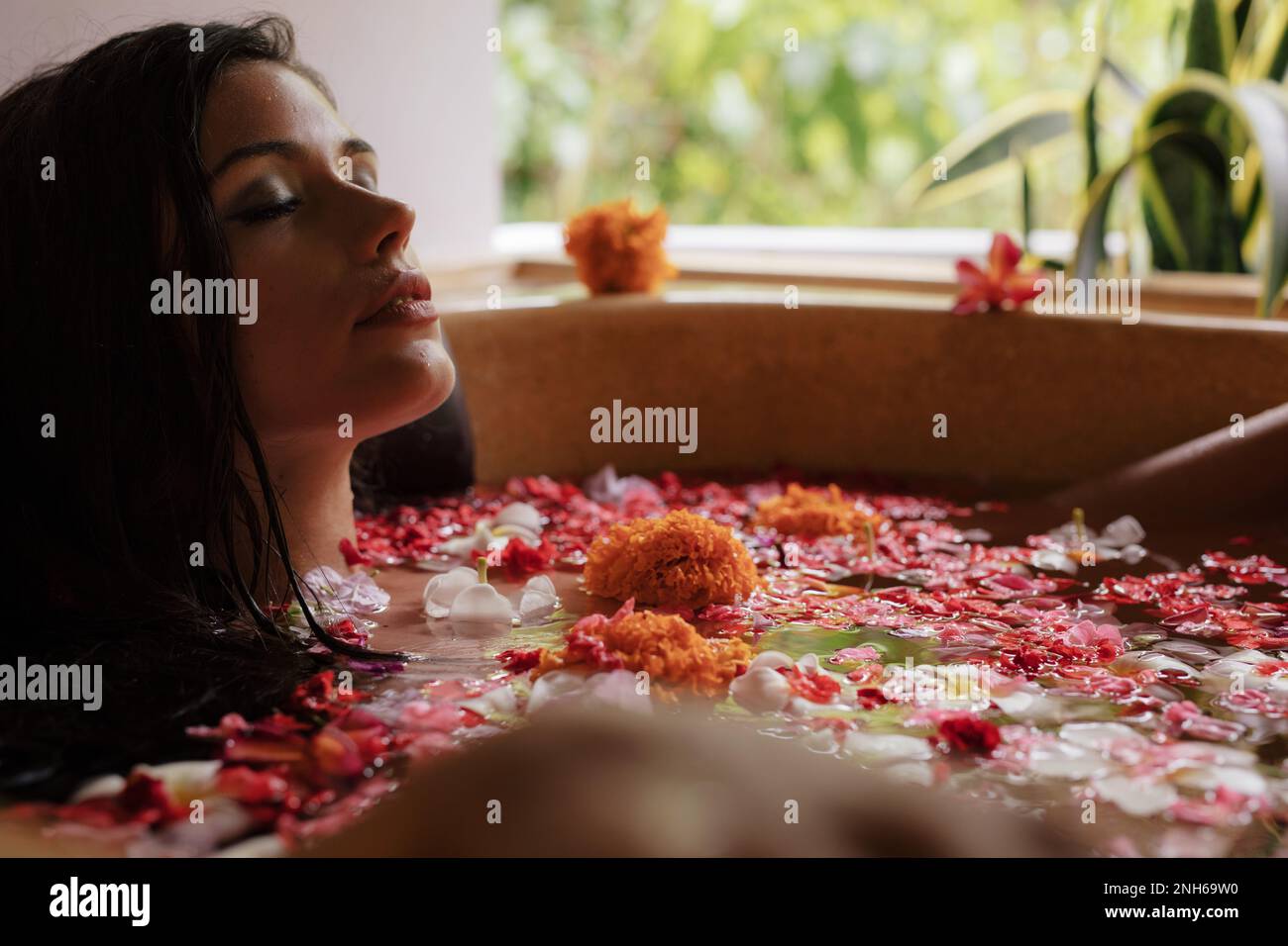Eine schöne junge Frau, die sich in der Badewanne mit Blütenblättern entspannt. Eine Frau, die ein Blumenbad in einem Spa hat. Stockfoto