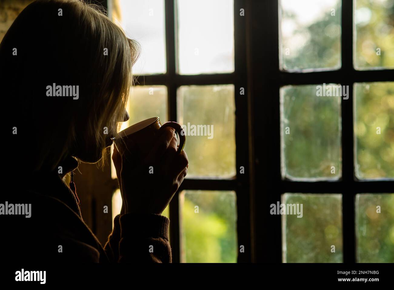 Dunkles Profil einer Frau mit einem Becher in der Hand am Fenster. Morgenkaffee. Stockfoto