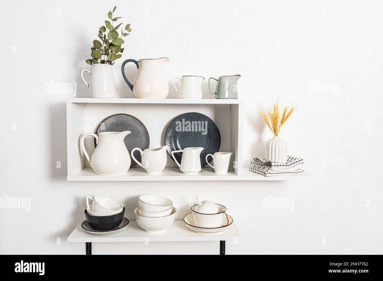 In den offenen Regalen in der Küche befinden sich viele verschiedene Kannen unter Wasser und Milch. Schüsseln und Teller. Wunderschöne weiße Innenansicht der Küchenregale Stockfoto