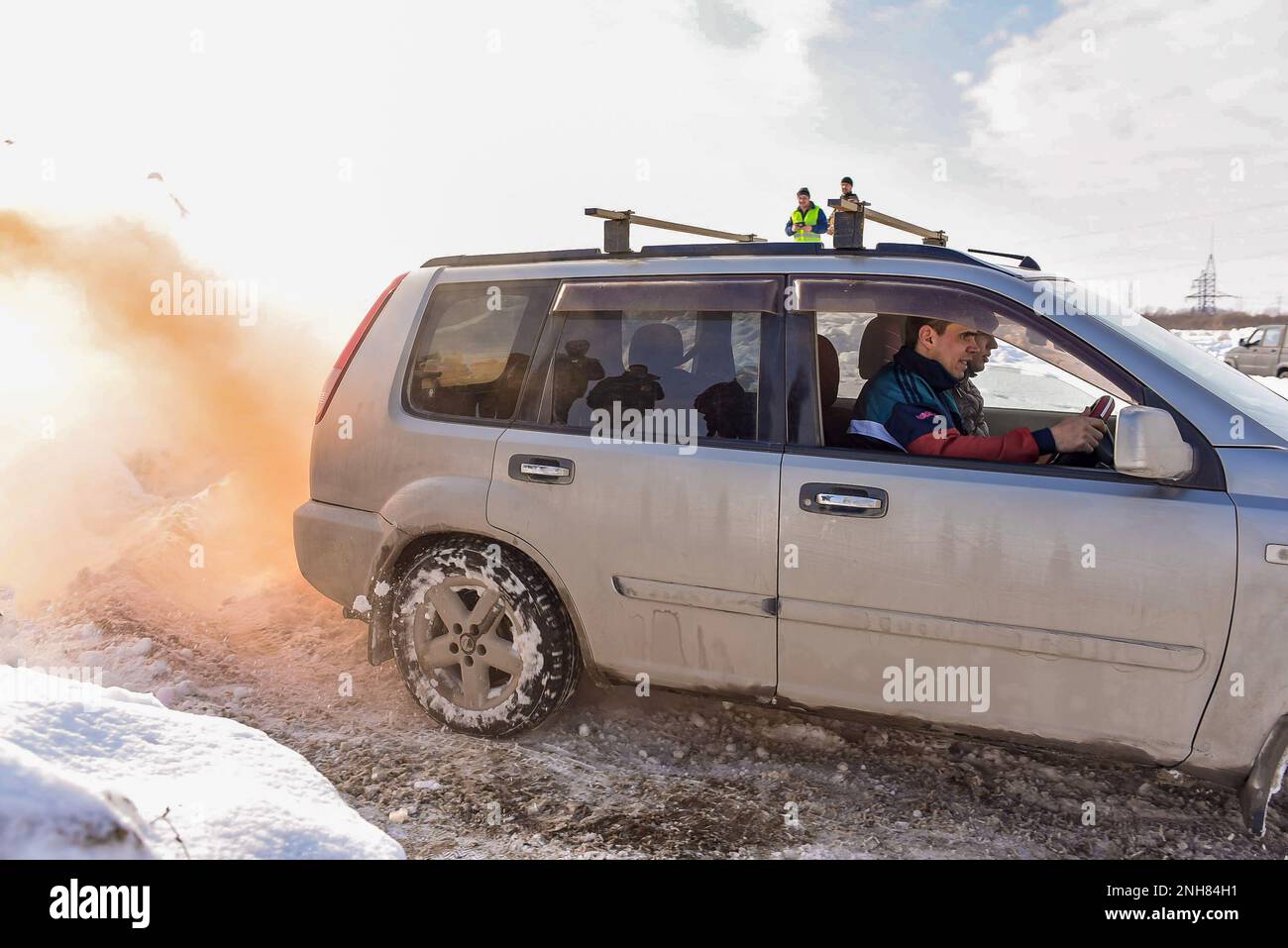 Offroad Crossover „Nissan X-Trail 4x4“ fährt im Winter schnell auf einer verschneiten Straße in orangefarbenem Rauch mit einem männlichen Fahrer in einem Trainingsanzug. Stockfoto