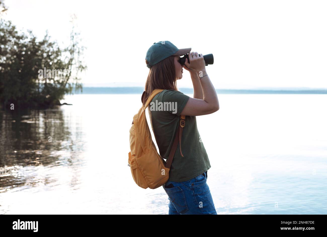 Junge Frau mit gelbem Rucksack, die durch ein Fernglas Vögel am See oder Fluss ansieht Vogelbeobachtung, Zoologie, Ökologie Forschung in der Natur, Beobachtung o Stockfoto