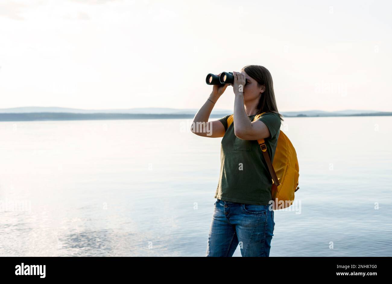Junge Frau mit gelbem Rucksack, die durch ein Fernglas Vögel am See oder Fluss ansieht Vogelbeobachtung, Zoologie, Ökologie Forschung in der Natur, Beobachtung o Stockfoto