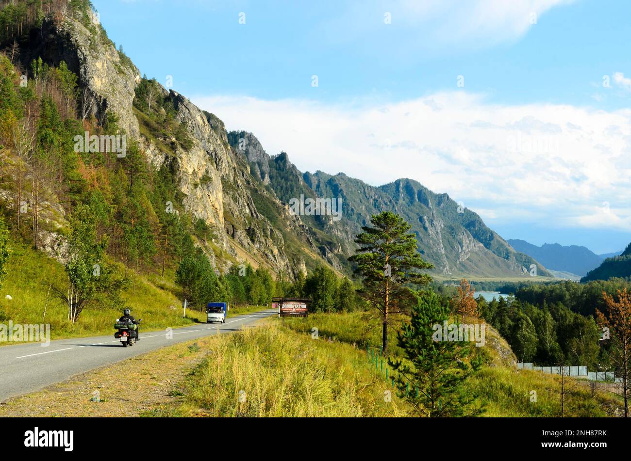 Ein Motorradfahrer und ein Auto, das auf einer Bergstraße am Hotel vorbeifährt, befinden sich neben einem Fluss im Altai Stockfoto