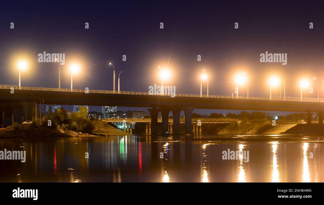 Nachtstraße mit Laternen, die von der Bogenbrücke in einen Fluss mit Reflexionen in Russland führt. Stockfoto