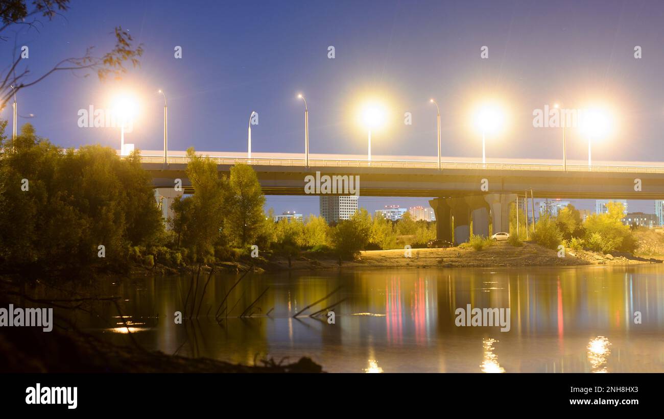 Nachtstraße mit Laternen, die von der Bogenbrücke in einen Fluss mit Reflexionen in Russland führt. Stockfoto