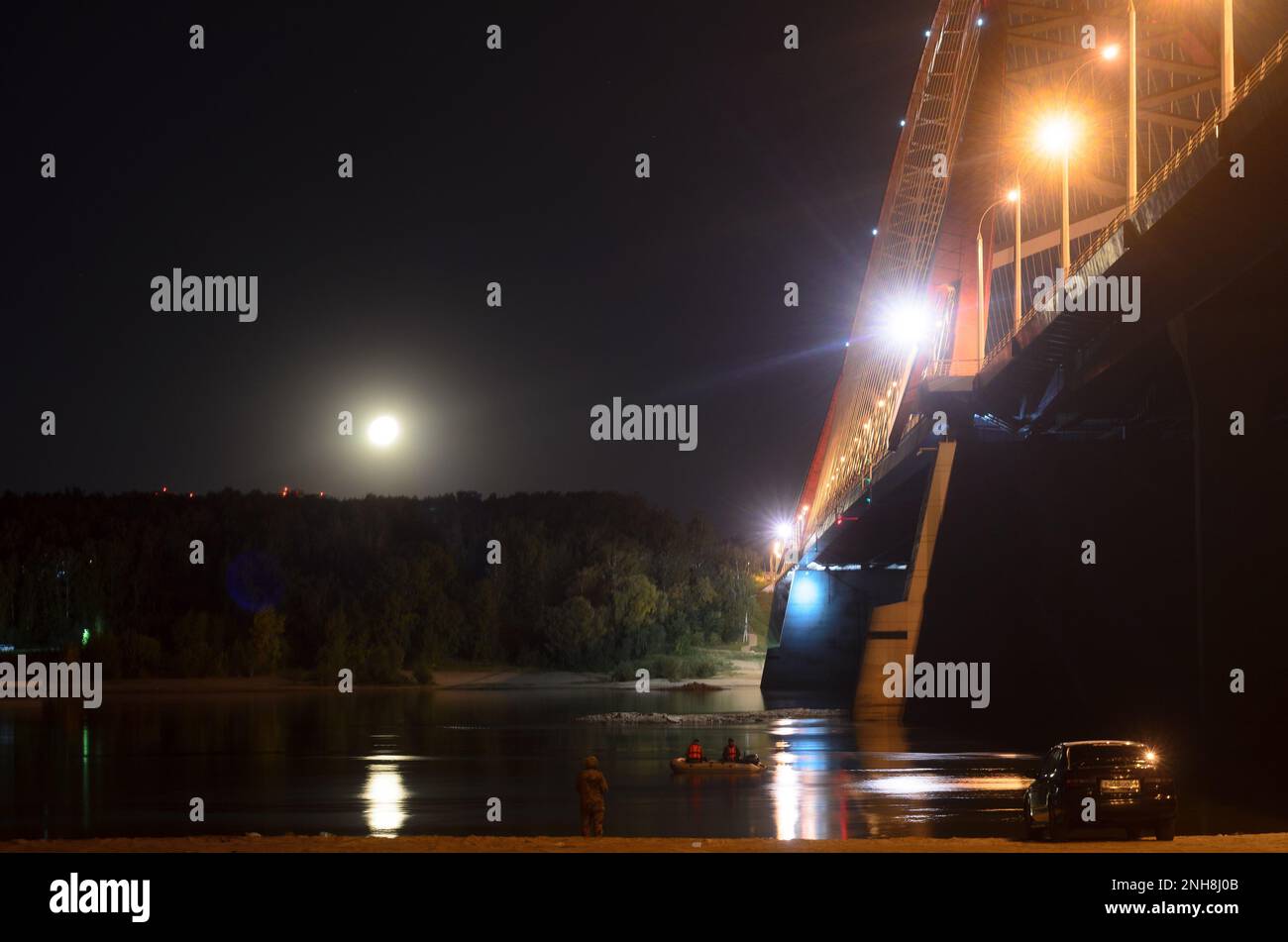 Zwei Männer fischen nachts in Booten unter einer großen leuchtenden Brücke am Ufer mit einem Auto Stockfoto