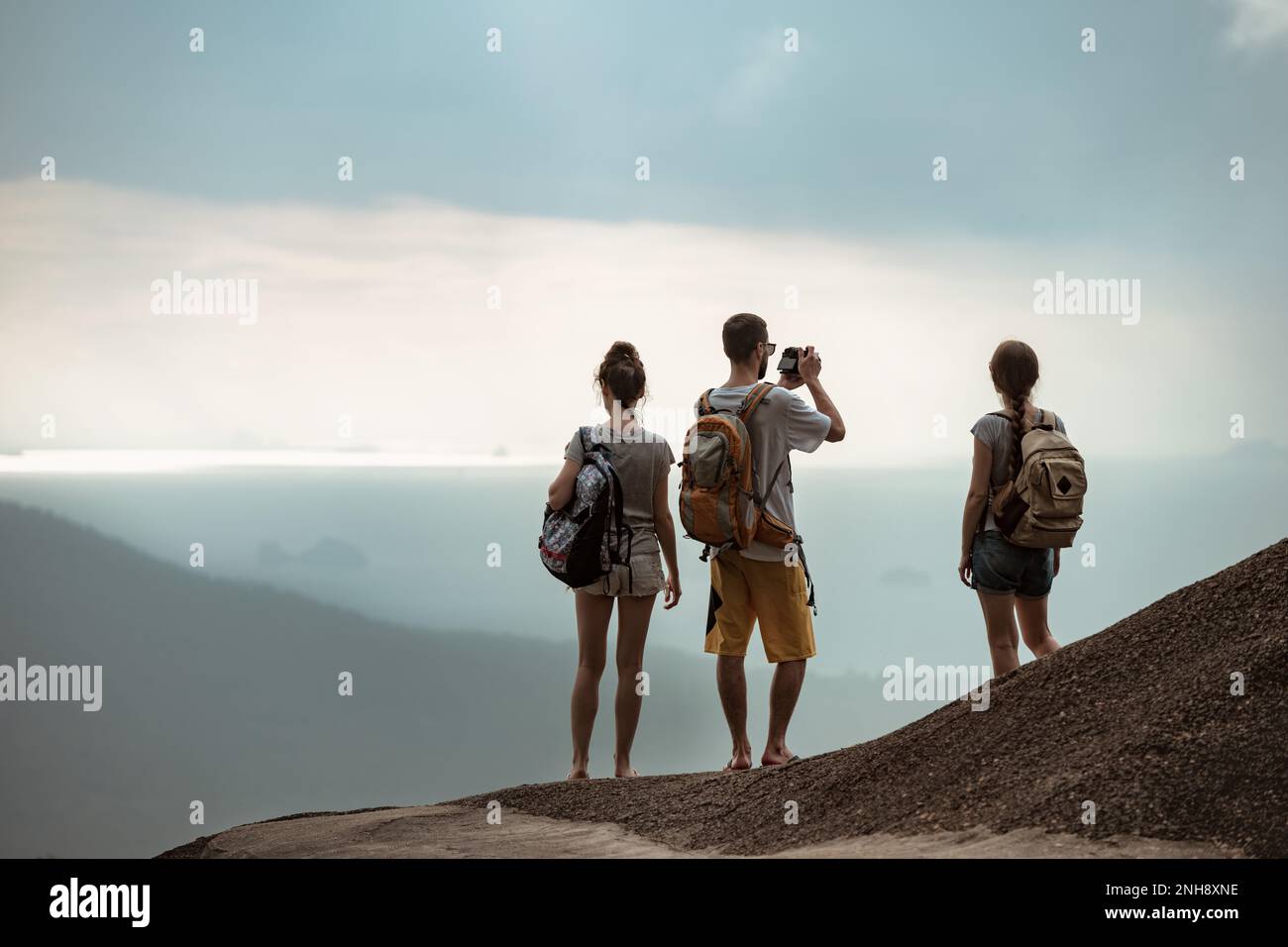 Drei junge Touristen mit Rucksäcken stehen auf dem Gipfel des Berges und blicken auf das Meer Stockfoto