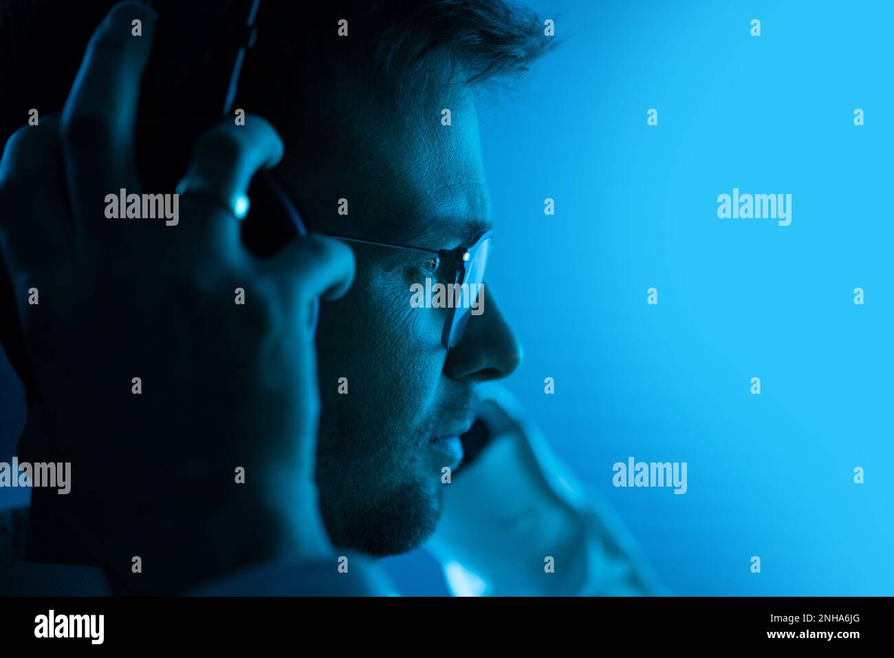 Kaukasischer Musikproduzent in seinem 30s, der mit professionellen Kopfhörern Musik hört. Unterhaltungsindustrie. Dunkelblau Beleuchteter Raum. Stockfoto