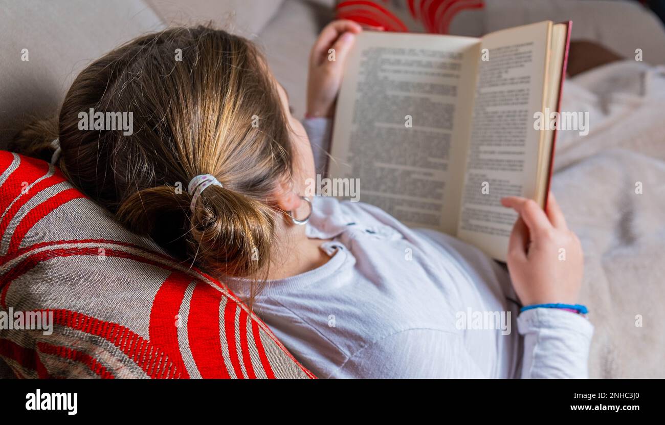 Ein Mädchen, das auf der Couch liegt und leise ein Buch liest. Weltbuchtag, Kinderleseaktion, Privatleben Stockfoto