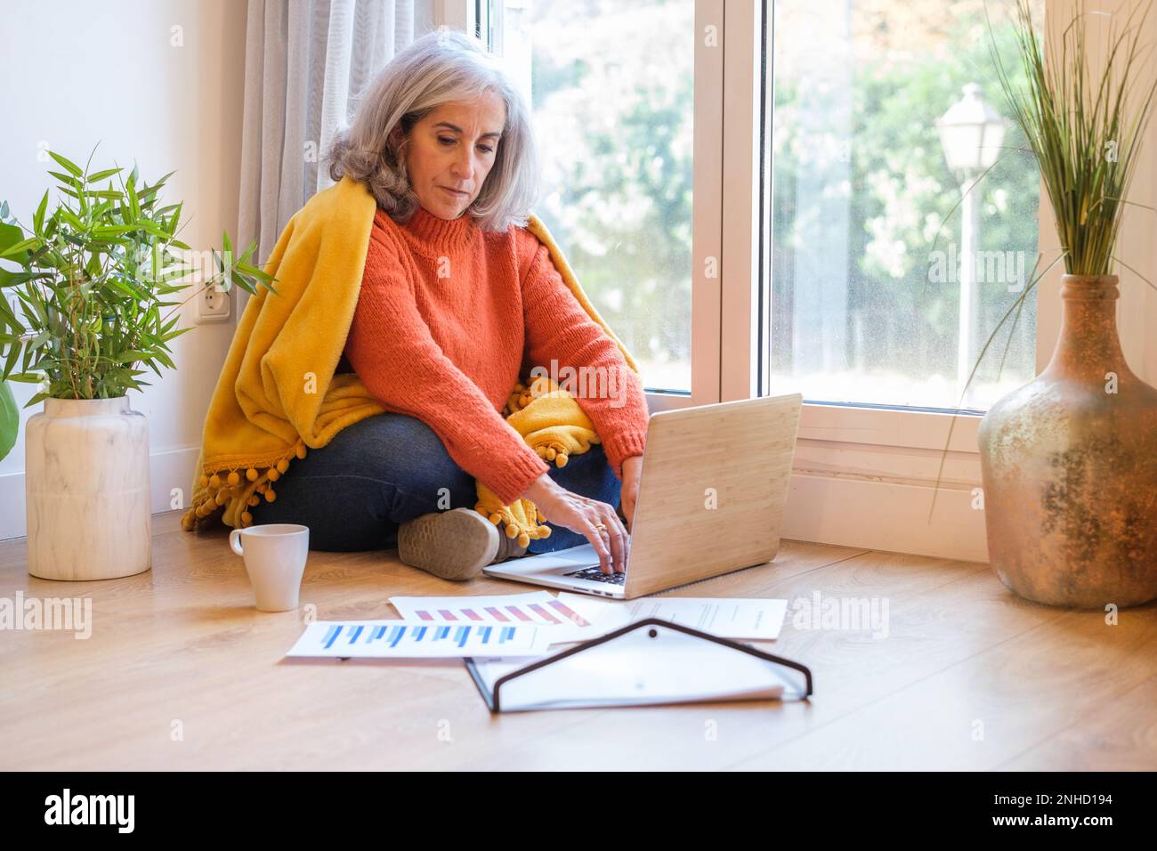 Seniorin mit weißen Haaren, die von zu Hause aus arbeitet, sitzt auf dem Boden am Fenster, mit Grafiken und dem Laptop. Konzept: Telearbeit, digitale Nomade, Stockfoto