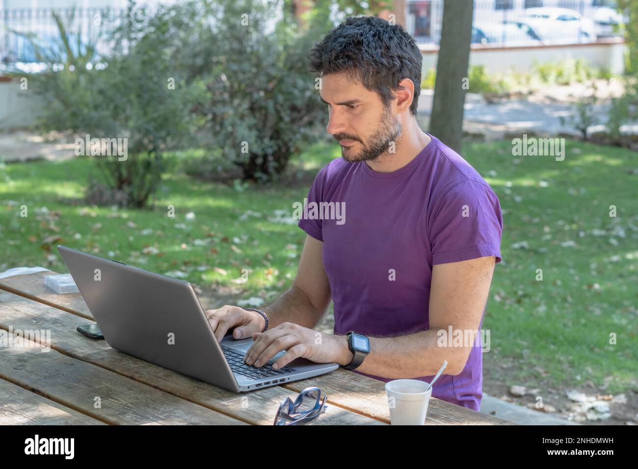 Junger Mann mit Bart und Frelance-T-Shirt, der im Park an seinem Computer arbeitet Stockfoto