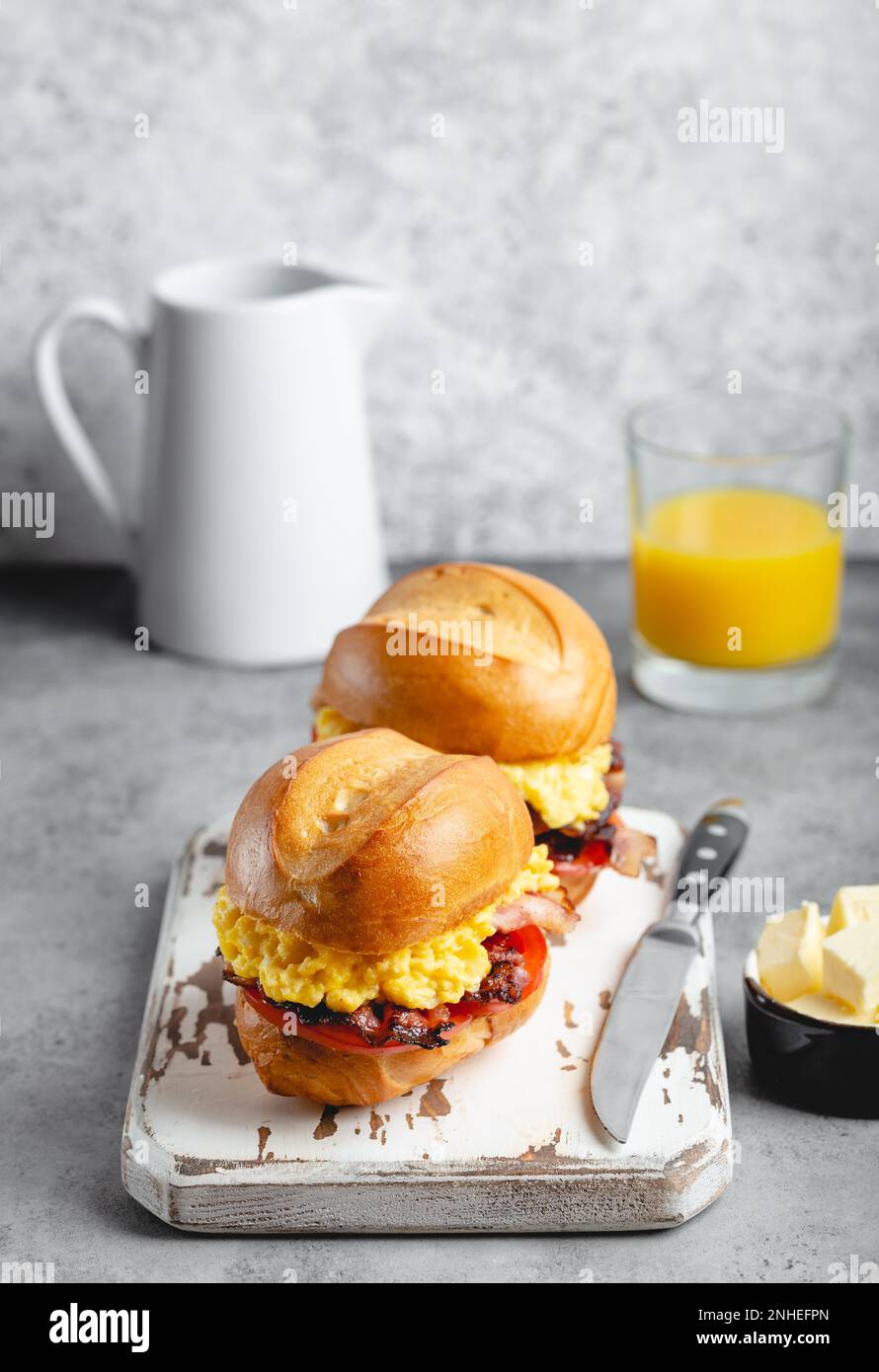 Frühstückssandwiches mit Rührei, Speck, Käse, Tomaten auf weißem Holzbrett, Glas mit frischem Orangensaft, weißer Hintergrund. Machen Stockfoto