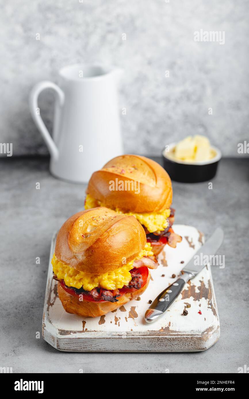 Frühstückssandwiches mit Rührei, Speck, Käse, Tomaten auf weißem Holzbrett, Glas mit frischem Orangensaft, weißer Hintergrund. Machen Stockfoto