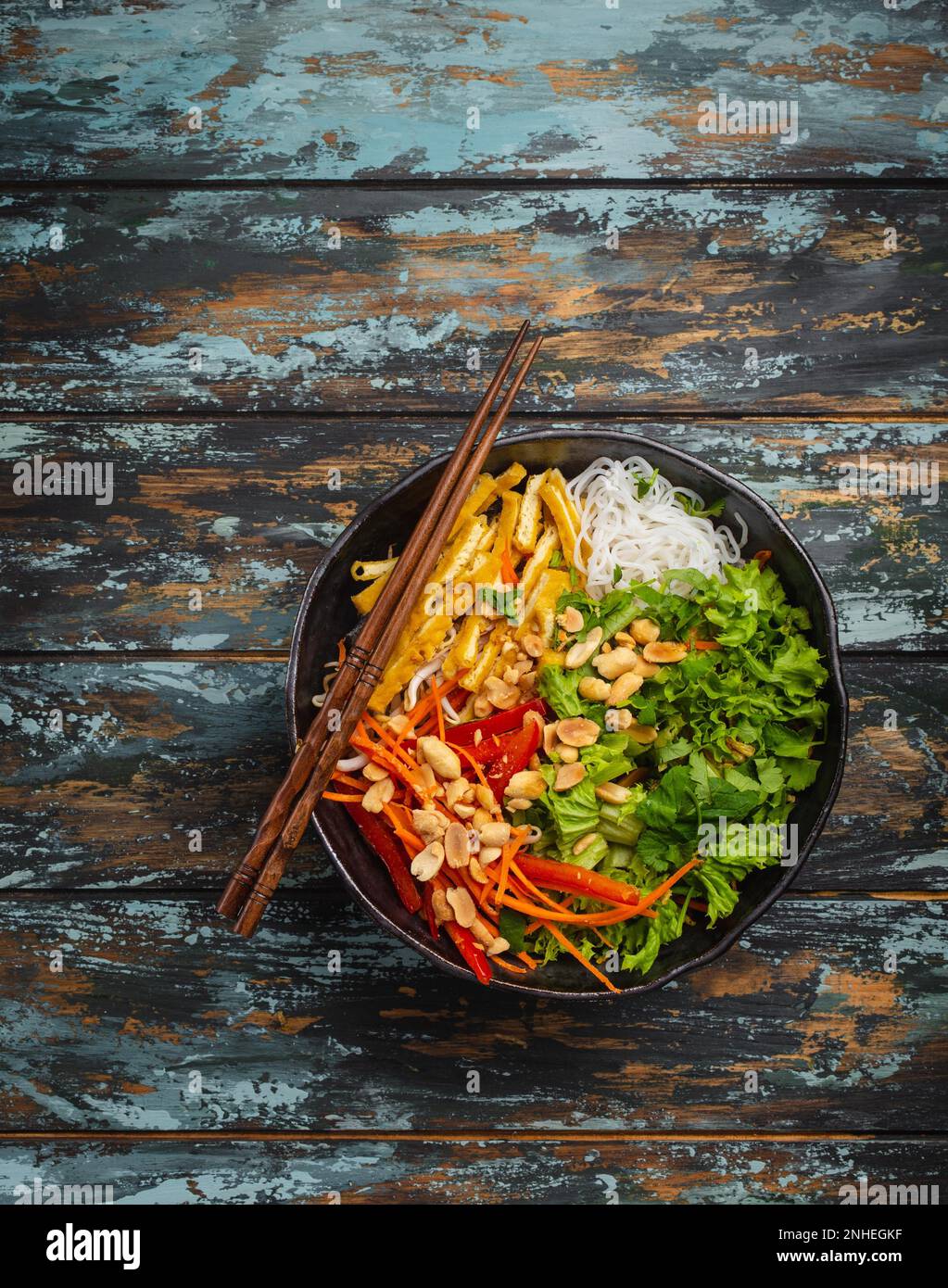 Nudelsalat im asiatischen, chinesischen oder vietnamesischen Stil mit frischem Gemüse, gebratenem Tofu und Erdnüssen, serviert in einer rustikalen Keramikschüssel auf farbenfrohem Holz Stockfoto