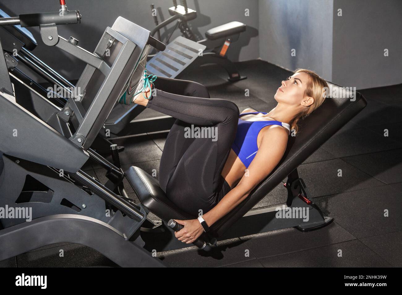 Seitliches Porträt einer starken Frau, die ein Fitnesstraining mit einer Beinverlängerung durchführt, eine Maschine mit Gewichten in einem Fitnessstudio schiebt und ein Sport-Top und eine Strumpfhose trägt. Indoor-Aufnahme. Stockfoto