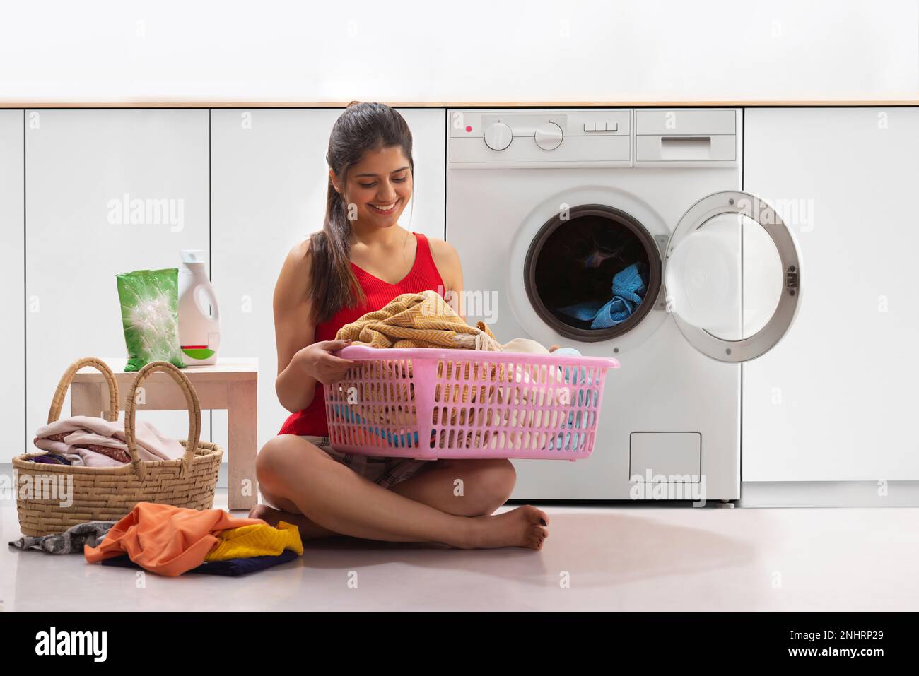 Lächelnde Frau, die vor der Waschmaschine sitzt, mit Wäschekorb Stockfoto