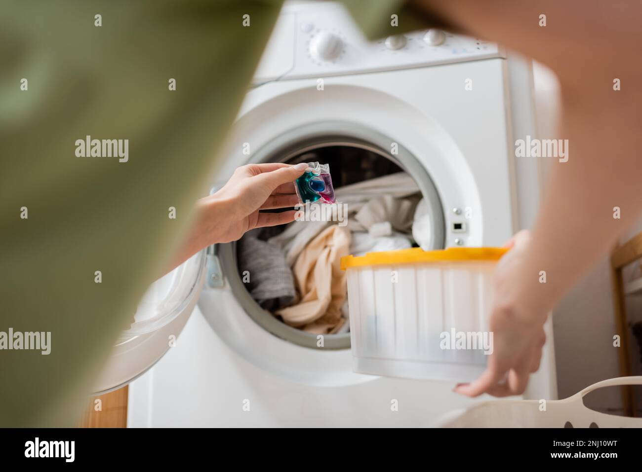 Zugeschnittene Ansicht einer Frau, die zu Hause in der Waschmaschine eine Flüssigwaschmittel-Kapsel in der Nähe von Kleidung hält, Stockbild Stockfoto