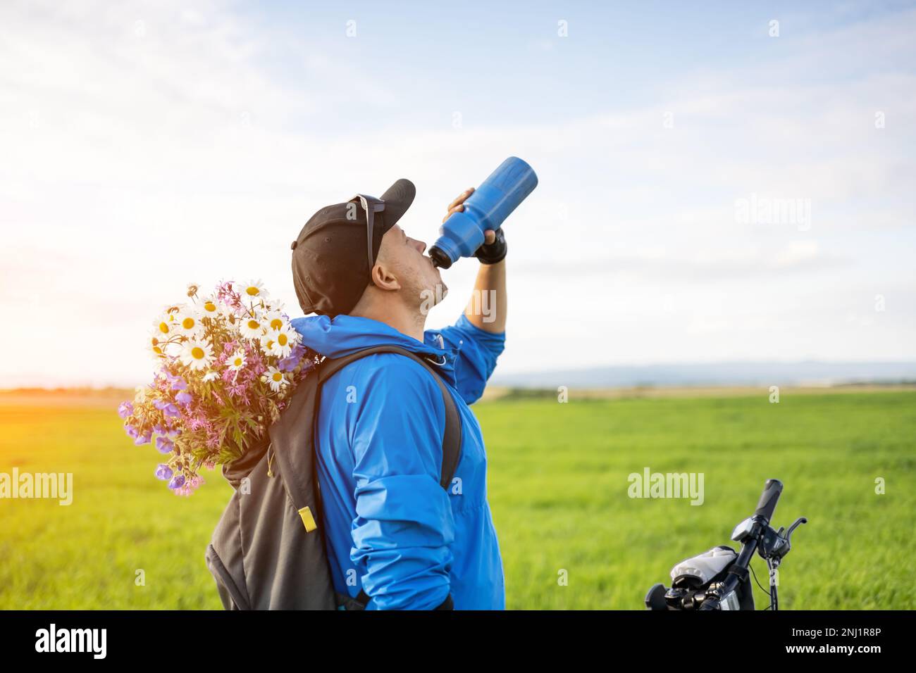 Mann mit Fahrrad trinkt Wasser im Frühlingsfeld. Ein Blumenstrauß im Biker-Rucksack Stockfoto