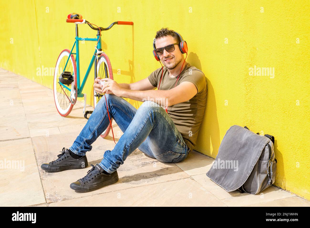 Rückansicht eines jungen Mannes, der auf dem Boden sitzt und sich an eine gelbe Wand lehnt und Musik über Kopfhörer hört, während er ein Mobiltelefon benutzt. Das hat er Stockfoto