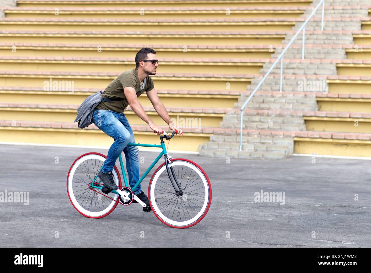 Ein junger Mann trägt eine Sonnenbrille und eine Schultertasche, während er mit dem Fixie Bike fährt. Stockfoto