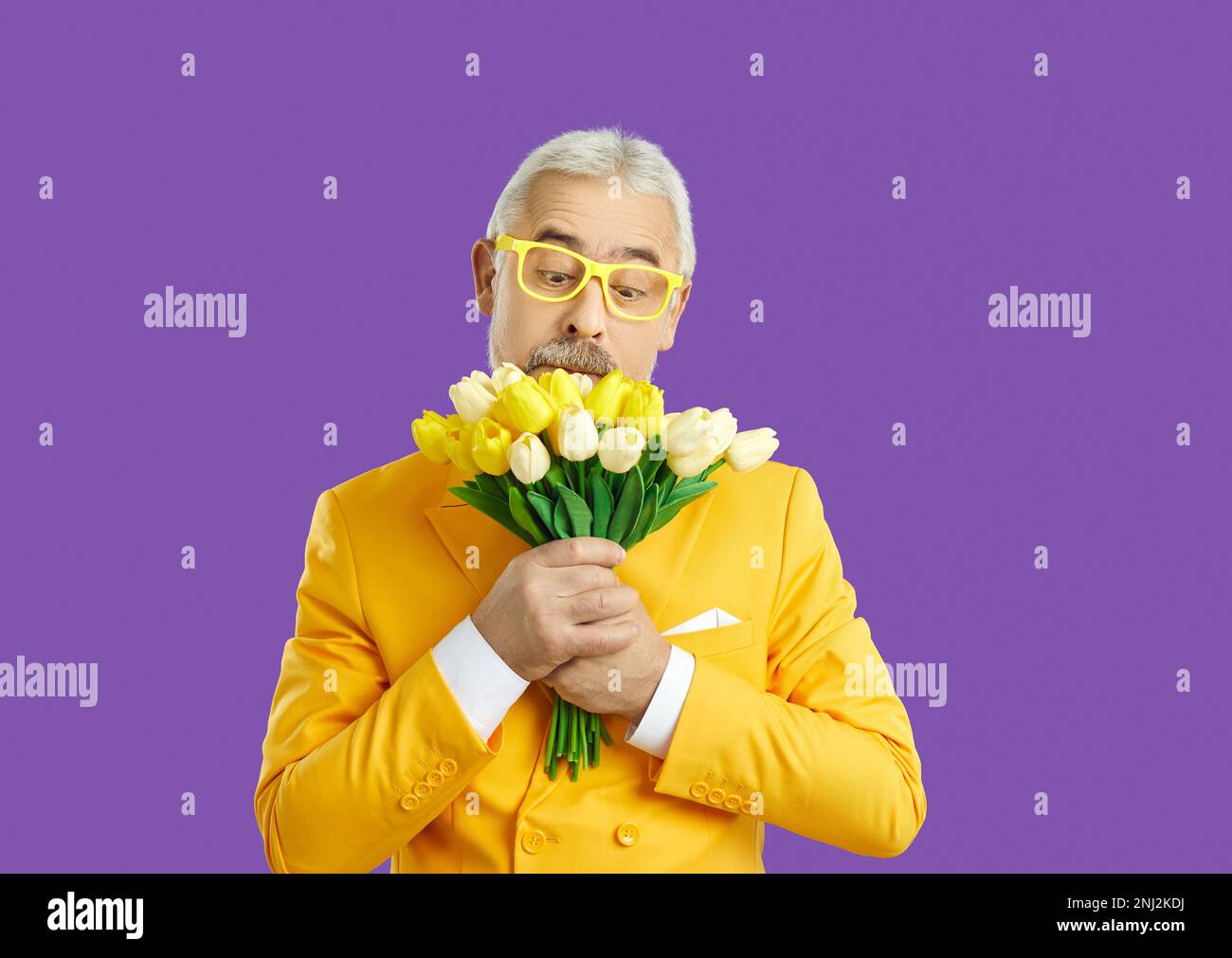 Ein erfahrener Mann mit komischem Gesichtsausdruck sieht sich einen Strauß Tulpen in seinen Händen an, auf lila Hintergrund. Stockfoto