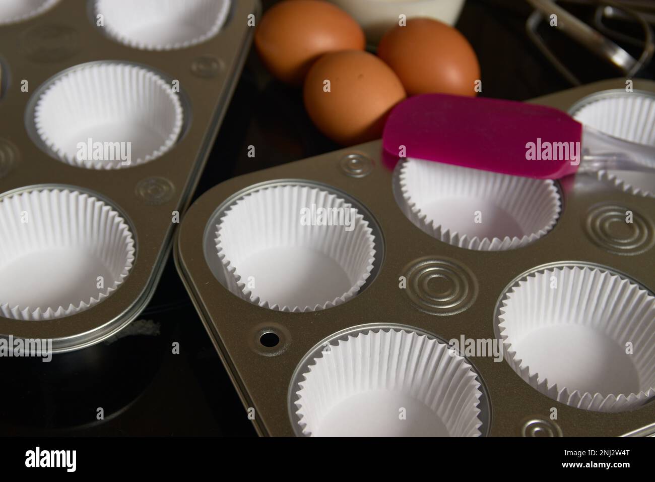 Nahaufnahme einiger Tabletts, die für die Zubereitung von Muffins vorbereitet wurden, zusammen mit braunen Eiern und ein paar Küchenzunge Stockfoto
