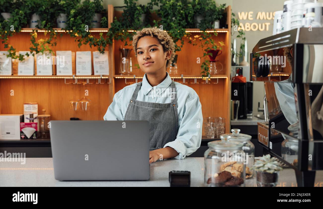 Junge Frau, die einen Coffee Shop besaß, mit einem Laptop am Tresen. Eine Barista in einer Schürze, die in einem Café arbeitet. Stockfoto