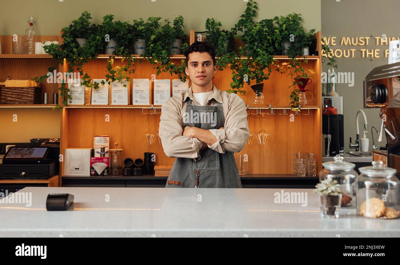 Ein junger selbstbewusster Barista, der beim Stehen an der Theke gerade in die Kamera schaut Stockfoto