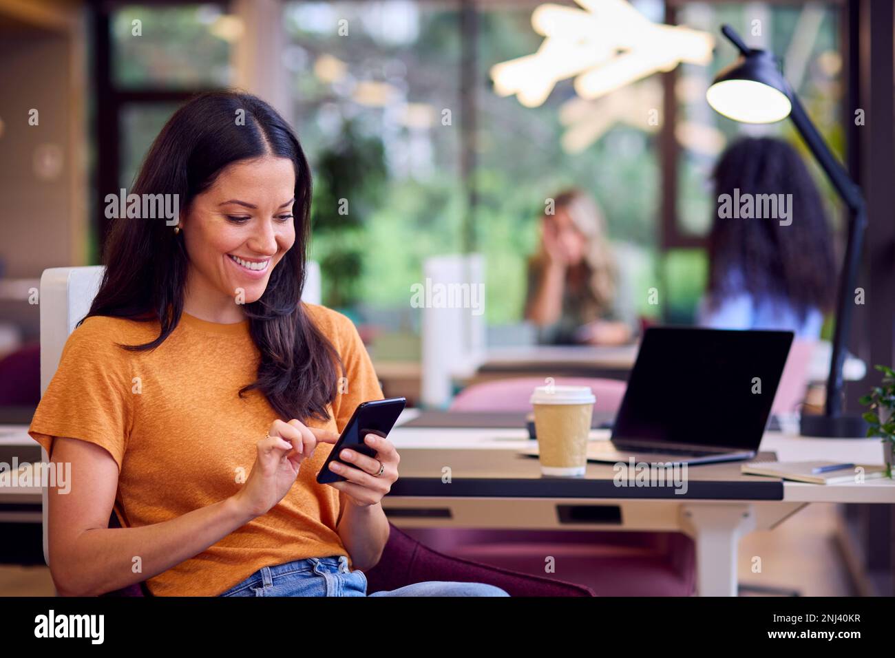 Eine Geschäftsfrau Sitzt Am Schreibtisch Im Büro Und Trinkt Mit Einem Schluck Und Schaut Auf Ein Mobiltelefon Stockfoto
