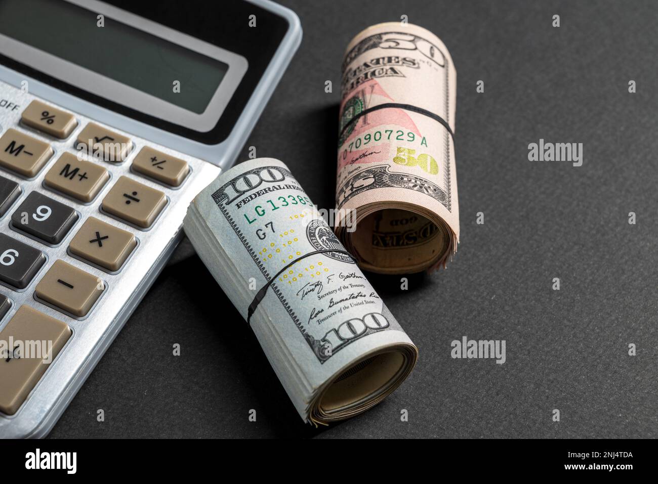 Rollen mit 50 und 100 US-Dollar stehen neben dem Rechner auf dunklem Hintergrund Stockfoto