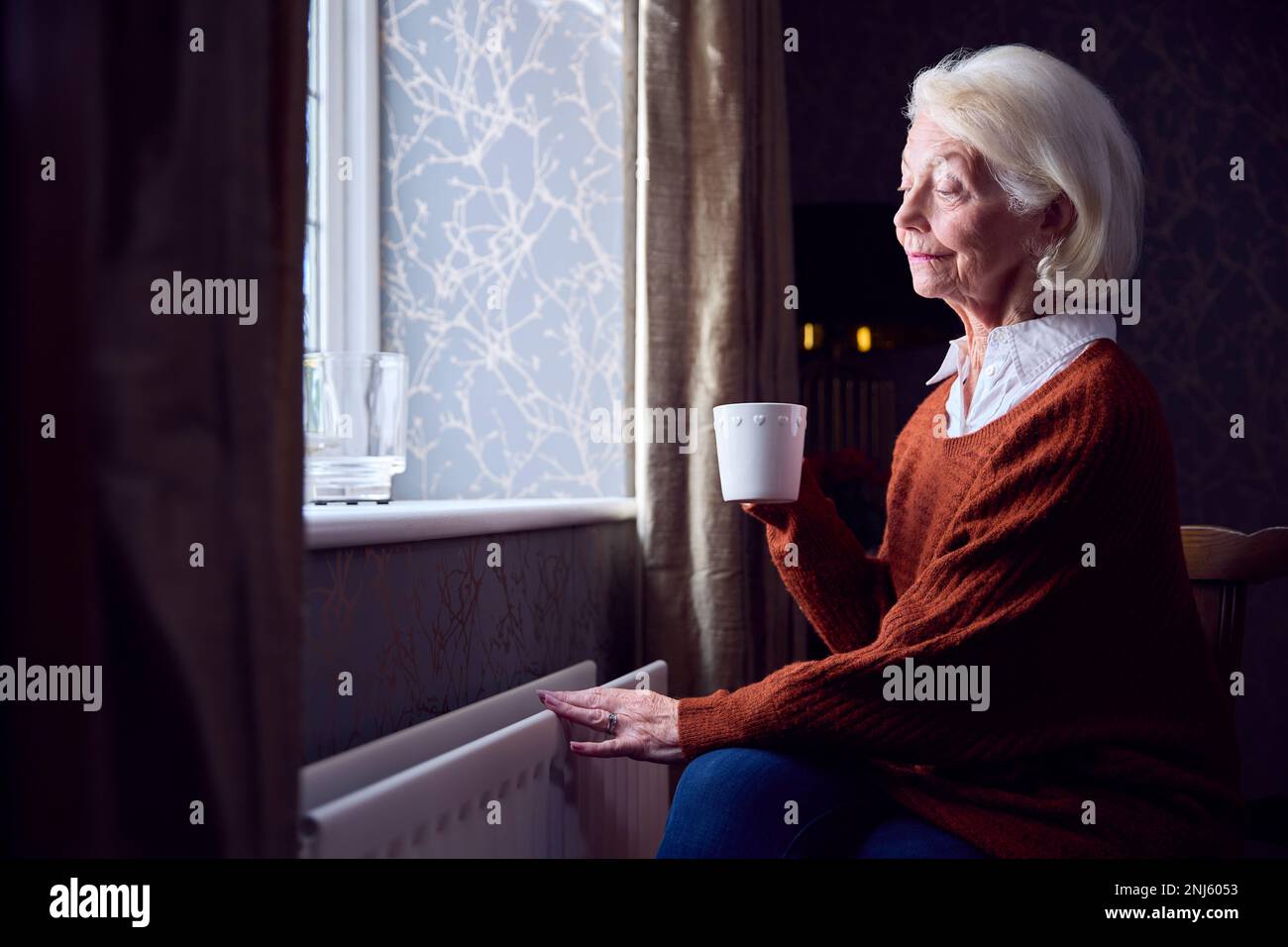 Seniorin Mit Heißem Getränk, Die Sich Bei Der Energiekrise "Kosten Des Lebens" Durch Heizkörper Warm Halten Will Stockfoto